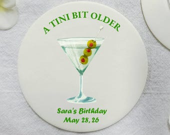 以下が含まれることがあります： 緑色の文字で「A TINI BIT OLDER」と書かれた白い丸いコースターと、オリーブの入った緑色のマティーニグラス。グラスの下には「Sara's Birthday May 28, 26」と書いてあります。