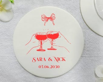 Può includere: Un sottobicchiere rotondo bianco con un'illustrazione rossa di due mani che brindano con calici di vino. Il sottobicchiere riporta i nomi "SARA & NICK" e la data "05.06.2030". Un calice e dei fiori sullo sfondo.