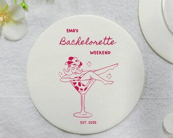 Può includere: Sottobicchiere rotondo bianco con testo rosa che recita "Ema's Bachelorette Weekend Est. 2025". Un'illustrazione di cartone animato di una donna in costume da bagno seduta in un bicchiere da martini si trova al centro del sottobicchiere.
