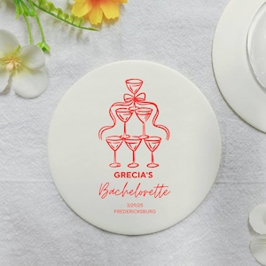 May include: Round white coaster with a red illustration of a champagne tower and the text "GRECIA'S Bachelorette 3/29/25 FREDERICKSBURG"