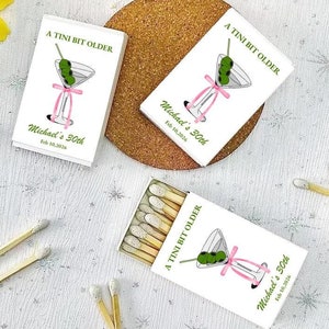 May include: Three white matchboxes with a martini glass design and the text "A Tini Bit Older" and "Michael's 30th" on the front. The matchboxes are on a white surface with some wooden matches scattered around.