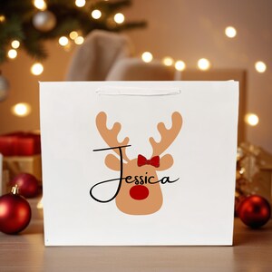 Puede incluir: Bolsa de regalo blanca con un diseño de reno, que incluye cuernos, una pajarita roja y una nariz roja. El nombre "Jessica" está escrito en cursiva debajo de los cuernos. La bolsa tiene un asa blanca, ideal para un regalo de Navidad.