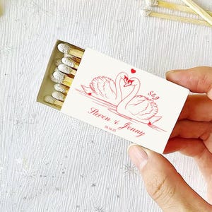 May include: A matchbox with a red swan design and the names "Steven & Jenny" printed on the front. The matchbox contains several wooden matches with white tips. The date "10.16.25" is also printed on the front.