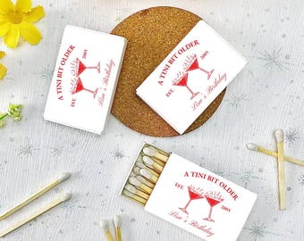 May include: A set of white matchboxes with a red design. The matchboxes have a red illustration of two martini glasses and the text "A TINI BIT OLDER EST. 2005 Lisa's Birthday".