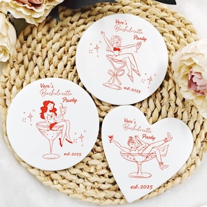 May include: Three white coasters with red illustrations and text. Two are round, one is heart-shaped. Each features a woman in a cocktail glass, with the text "Vem's Bachelorette Party" and "est. 2025". The coasters are on a woven mat.