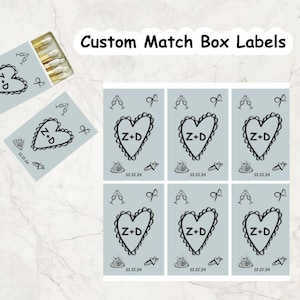 May include: Custom match box labels with a light blue background. Each label features a black heart with the initials "Z+D" and the date "12.15.24". The labels also include small illustrations of champagne glasses, a cake, and a bow.