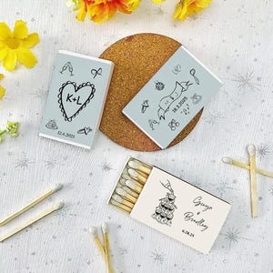 May include: Three matchboxes with custom designs. Two are light blue with black illustrations and dates. The third is off-white with a wedding dress illustration and names. Matches are visible in one box.