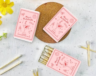 May include: Pink matchboxes with the text "Olivia's Bachelorette" and a cocktail illustration. The matchboxes are decorated with bows, hearts, and a disco ball. The matches are visible inside one box.