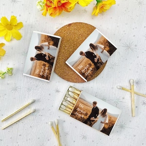 May include: A set of matchboxes with a photo of a couple on the front. The matchboxes are white with the text "The Perfect Match" printed on them. The matchboxes are on a white surface with some yellow flowers in the background.