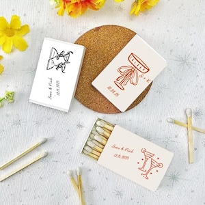 Peut inclure: Trois bo&icirc;tes d'allumettes blanches avec des illustrations noires et rouges de coupes de champagne. Les bo&icirc;tes d'allumettes portent l'inscription "Sara & Nick 12.15.2025" et "S + K 10.24.25".