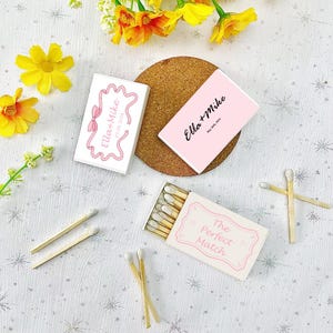 May include: Two white matchboxes with pink accents. One box has the text "Ella + Mike" and the date "May 30, 2006". The other box has the text "Ella + Mike" and the date "01.06.2026". A third matchbox has the text "The Perfect Match" in pink lettering.  The matchboxes are surrounded by yellow flowers and matchsticks.