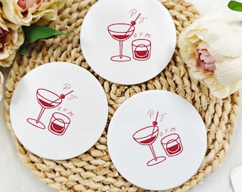 May include: Three white ceramic coasters with a red cocktail and whiskey glass design. Each coaster has the initials "PJ" and the date "12.11.25". The coasters are arranged on a woven mat, with flowers in the background.
