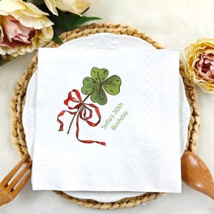 Puede incluir: Servilleta cuadrada blanca con una ilustración de trébol de cuatro hojas verde y una cinta roja. El texto "Sofia's 30th Birthday" está impreso en verde. La servilleta está sobre un plato blanco y un salvamanteles tejido.
