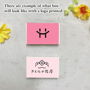May include: Two pink boxes with different logos printed on them. The top box has a simple smiley face logo. The bottom box has a frog with a flower on its head and the text "カエルの花屋" in Japanese.