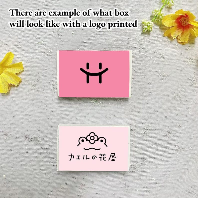 May include: Two pink boxes with different logos printed on them. The top box has a simple smiley face logo. The bottom box has a frog with a flower on its head and the text "カエルの花屋" in Japanese.