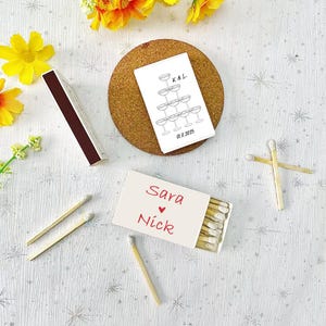 Peut inclure: Bo&icirc;te d'allumettes blanche avec les noms Sara et Nick &eacute;crits en rouge avec un symbole de c&oelig;ur entre eux. La bo&icirc;te d'allumettes est sur une surface blanche avec plusieurs allumettes en bois &eacute;parpill&eacute;es autour. Une autre bo&icirc;te d'allumettes blanche avec un dessin d'une tour de fl&ucirc;tes &agrave; champagne et le texte "K&L 01.11.2025" est sur un sous-verre en li&egrave;ge.