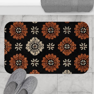 Puede incluir: Una alfombra de baño rectangular con un fondo negro y un patrón repetido de diseños florales naranjas y blancos. La alfombra está colocada sobre un suelo embaldosado, con un par de zapatillas grises visibles en la parte inferior.