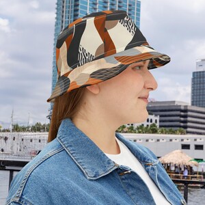 Könnte beinhalten: Ein Bucket Hat mit einem geometrischen Muster in Braun-, Schwarz-, Beige- und Grautönen. Der Hut hat eine breite Krempe und wird von einer Person mit einer Jeansjacke getragen. Das Design des Hutes zeigt abstrakte Formen.