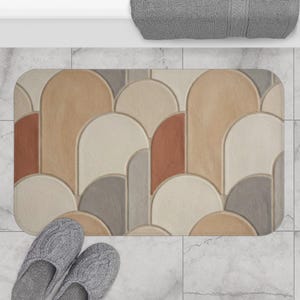 May include: A rectangular bath mat with a geometric pattern in shades of beige, brown, gray, and rust. The mat features a repeating arched design. Gray knit slippers are at the bottom of the mat.