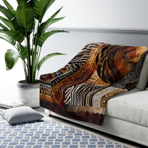 Może przedstawiać: Koc z wzorem inspirowanym Afryką, narzucony na białą sofę. Koc ma patchwork wzorów w odcieniach brązu, czerni i beżu, w tym paski zebry i kształty geometryczne. Duża roślina doniczkowa znajduje się w tle.