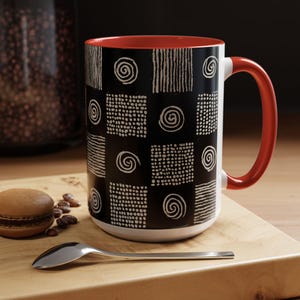 Może przedstawiać: Ceramiczny kubek z czarną powierzchnią zewnętrzną ozdobioną powtarzającym się wzorem białych spirali, kropkowanych kwadratów i kwadratów w paski. Wnętrze i uchwyt kubka mają jednolity, jasnoczerwony kolor, a podstawa i krawędź są białe.