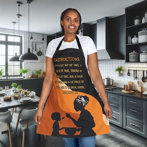 Puede incluir: Delantal negro con un gráfico naranja y negro de una madre y un niño cocinando, con el texto "INSTRUCTIONS: ADD A LIL' BIT OF THIS, A PINCH OF THAT...THEN TASTE IT!" El delantal tiene una correa negra para el cuello y lo lleva una persona con una camisa blanca.