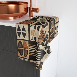 May include: A decorative hand towel with a geometric pattern in black, tan, and brown. The towel features various shapes and designs, including triangles, rectangles, and circles. The towel is draped over a white countertop.