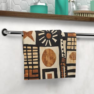 Puede incluir: Una toalla de mano estampada con un diseño geométrico en negro, beige y marrón. El diseño incluye formas como cuadrados, círculos y un sol. La toalla está colgada de una barra cromada.
