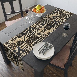 May include: A black and beige patterned table runner with geometric designs, displayed on a dark wooden table. The table is set with white plates, silverware, wine glasses, and a bowl of apples, creating a dining setting.