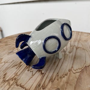 May include: A ceramic rocket ship planter or vessel featuring a light gray crackle glaze finish. The object has deep blue accents on the fins, the base, and two circular porthole-like rings on the side. It is shaped like a stylized space rocket.