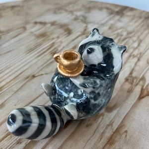 May include: A small ceramic raccoon figurine with a glossy black and white mottled finish, featuring a long striped tail and holding a miniature metallic gold-colored teacup and saucer in its front paws while in a seated position.