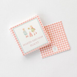 May include: Square stationery with a pink and white gingham pattern border. The center features a light blue border with a watercolor illustration of children and the text "MAREN AND SAM ELLIOTT". A matching card is next to it.
