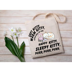 Puede incluir: Un bolso tote beige con una ilustración de gato y texto. El gato es blanco con manchas naranjas, durmiendo sobre libros. El texto incluye "Soft Kitty, Warm Kitty, Little Ball of Fur" y "Happy Kitty, Sleepy Kitty, Purr, Purr, Purr."