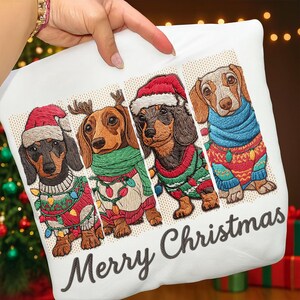Pode incluir: Fronha branca com um bordado de quatro dachshunds vestidos para o Natal. Cada cão usa uma roupa festiva, incluindo gorros de Papai Noel e suéteres. A frase "Merry Christmas" está escrita abaixo.