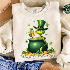 May include: Cream-colored sweatshirt with a St. Patrick's Day graphic. The design features a green character in a leprechaun hat holding a beer mug over a pot of gold. Green shamrocks and the text "Happy St. Patrick's Day" complete the festive look.
