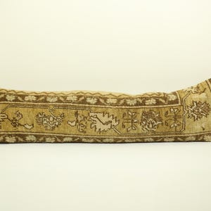 May include: A long, rectangular pillow with a brown and cream patterned fabric. The pillow is made from a woven fabric with a traditional design.