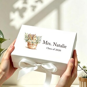 May include: A white rectangular gift box featuring a watercolor illustration of a pot filled with pencils and flowers. The text "Mrs. Natalie" and "Class of 2026" is printed in black. A white satin ribbon is tied in a bow at the front of the box.
