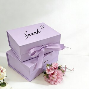 May include: Two stacked lavender gift boxes with a matching ribbon and bow. The top box is inscribed with the name "Sarah" and a heart symbol. Pink flowers are arranged near the boxes.