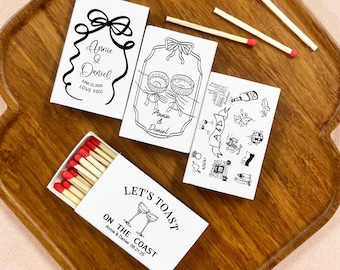 May include: A wooden serving tray holds several white matchboxes with black text and illustrations. One box is open, revealing red-tipped matches. The text includes "Annie & Daniel LOVE YOU" and "LET'S TOAST ON THE COAST Annie & Daniel 06.21.25".