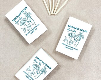 May include: White matchboxes with a teal beach-themed design and the text "Rosemary Beach Bach Club" and "Jessica's Bachelorette August 2023". Five matches with red tips are arranged near the matchboxes. The items are on a neutral surface.