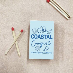 May include: A light blue matchbox with the words "COASTAL Cowgirl" and "KATE'S BACHELORETTE" printed on the front. The matchbox is decorated with illustrations of a cowboy hat, seashell, and horseshoe. Three matches with red tips are next to the matchbox.