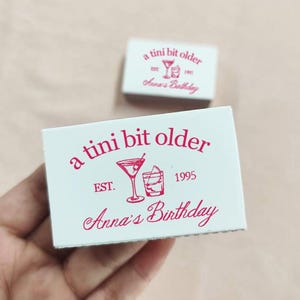 May include: A white matchbox with pink text that reads "a tini bit older est. 1995 Anna's Birthday". The matchbox features a pink illustration of a martini glass and a rocks glass.