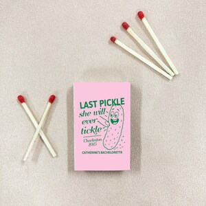 May include: A pink matchbox with the text "LAST PICKLE she will ever tickle" and a cartoon pickle illustration. The matchbox is accompanied by several white matches with red tips, arranged on a light pink surface. "Charleston 2025" and "CATHERINE'S BACHELORETTR" are also printed on the matchbox.