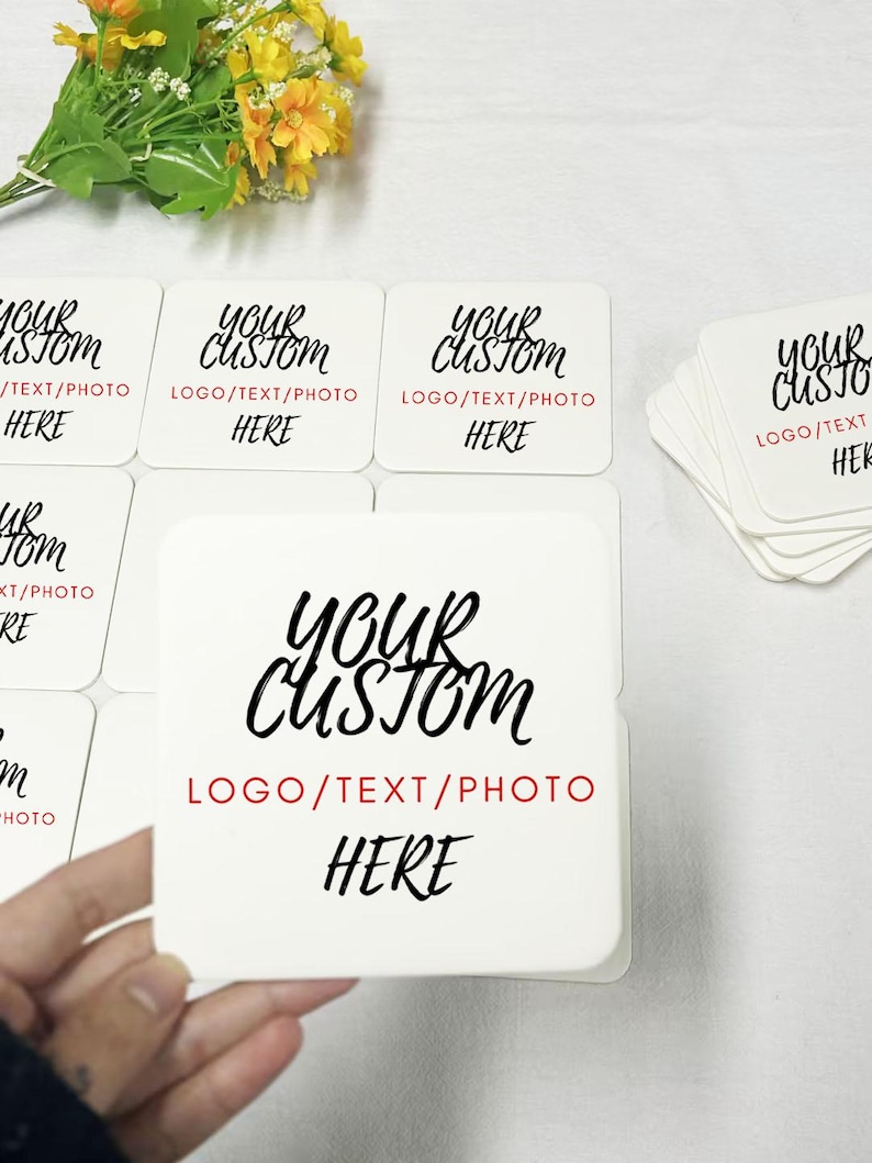 May include: A stack of white square coasters with the text "Your Custom Logo/Text/Photo Here" printed in black. The text is in a handwritten font. The coasters are on a white surface.