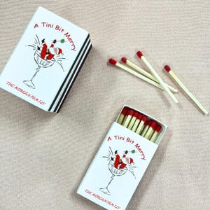 May include: White matchboxes with black striped edges and the text "A Tini Bit Merry" and "The Morgan Family". The boxes feature a Santa Claus illustration in a martini glass. Several loose matches with red tips are scattered nearby.