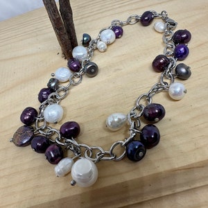 May include: A silver chain bracelet adorned with a mix of freshwater pearls. The pearls vary in size and color, including white, deep purple, and dark gray. The bracelet is displayed on a light wooden surface.