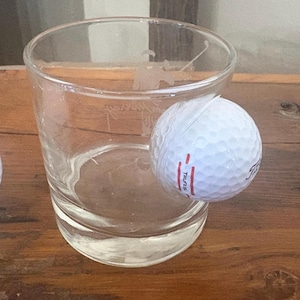 May include: A clear glass tumbler with a golf-themed etching and a white golf ball. The golf ball has red markings and the word "TruFeel" printed on it. The glass is on a wooden surface.