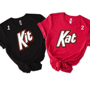 May include: Two short-sleeved t-shirts, one black and one red. The black shirt has the word "Kit" in white with a red and brown border. The red shirt has the word "Kat" in white with a red and brown border. Both shirts are on a white background.