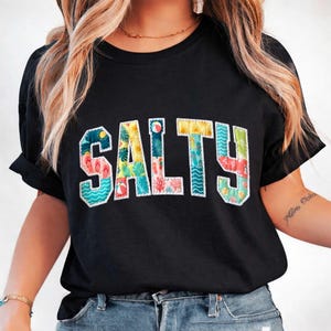 May include: Black t-shirt with the word "SALTY" in colorful, beach-themed letters. The letters are filled with images of flip-flops, palm trees, and other summer elements. The shirt has short sleeves and a crew neck.