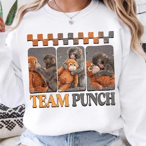 Puede incluir: Sudadera blanca con un gráfico de tres monos abrazando a un mono de peluche. El texto "TEAM PUNCH" está impreso en naranja y gris. La parte superior del gráfico tiene un patrón repetitivo de rectángulos naranjas y grises.